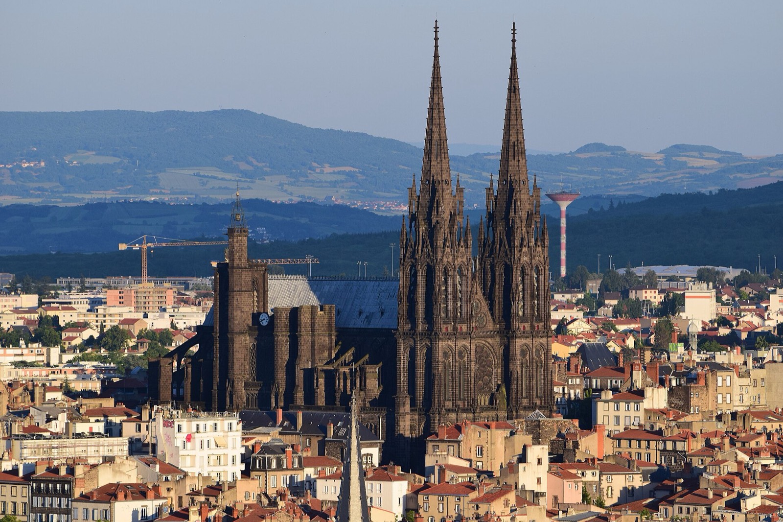 The dark twin spires of Clermont-Ferrand cathedral rising above the red-tiled roofs of the old town in evening light, with wooded hills behind