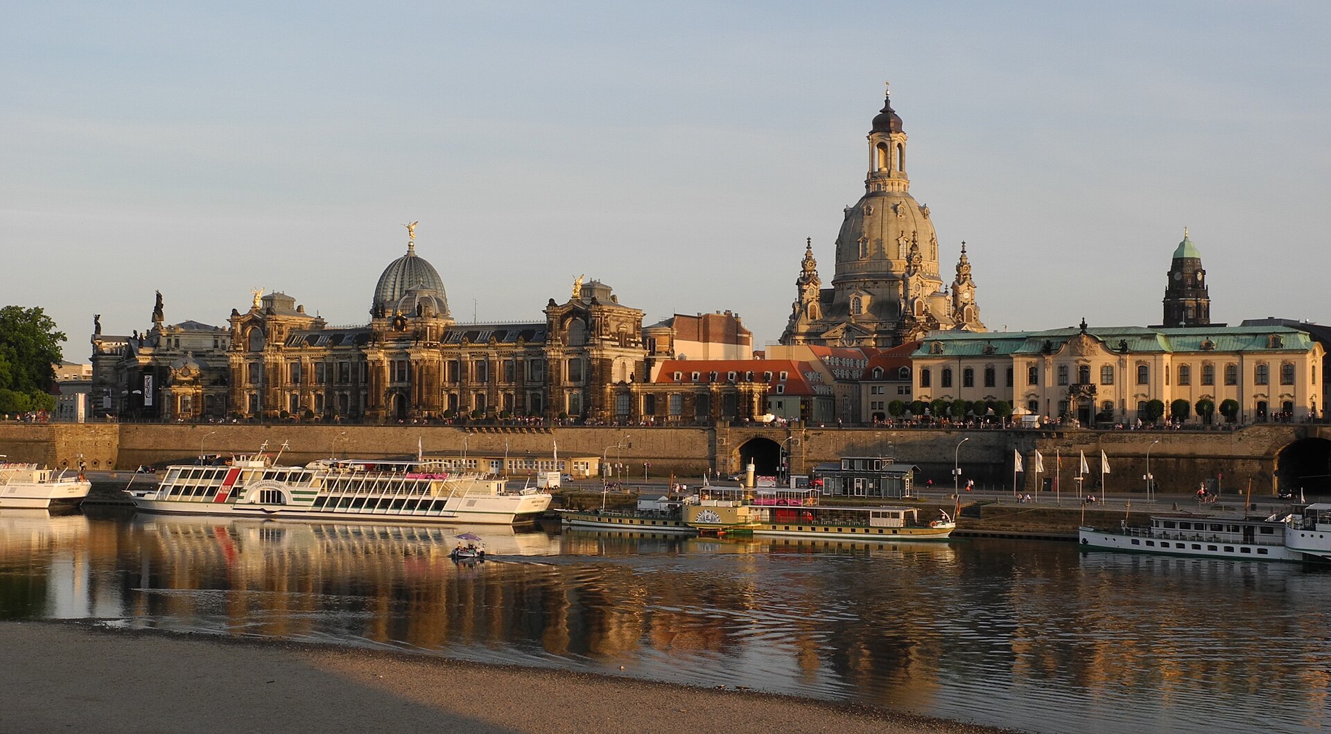 Dresden’s old town seen across the Elbe in warm evening light: the stone dome of the Frauenkirche above Brühl’s Terrace, the glass dome of the art academy to the left and excursion boats moored along the embankment