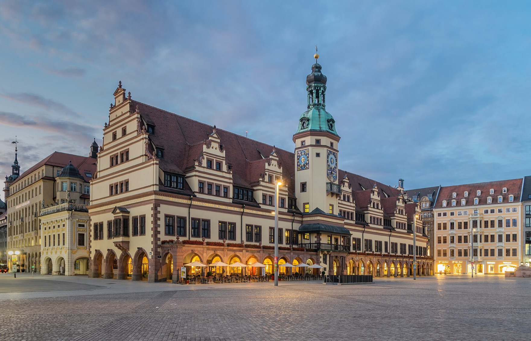 The Renaissance Old Town Hall on Leipzig’s market square at first light, its arcades lit from within and the clock tower with a green copper cap against a blue-grey sky