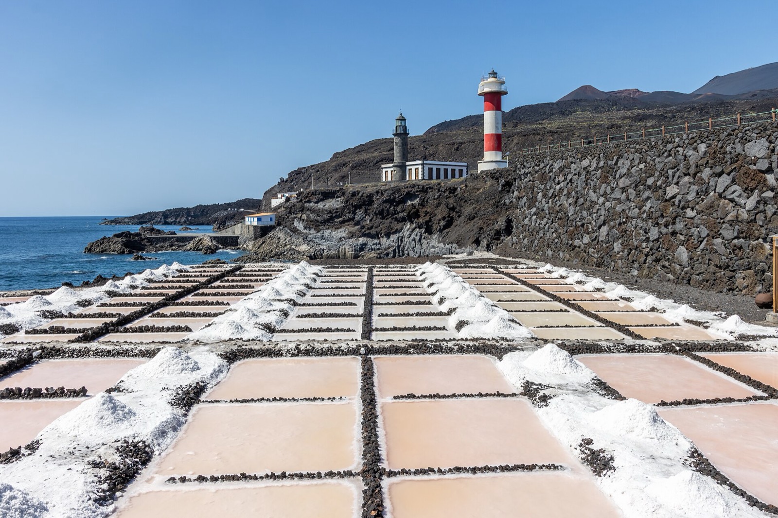 The salt pans of Fuencaliente on La Palma: pale pink brine pools and heaps of white salt, with two lighthouses on dark lava above and the Atlantic to the left