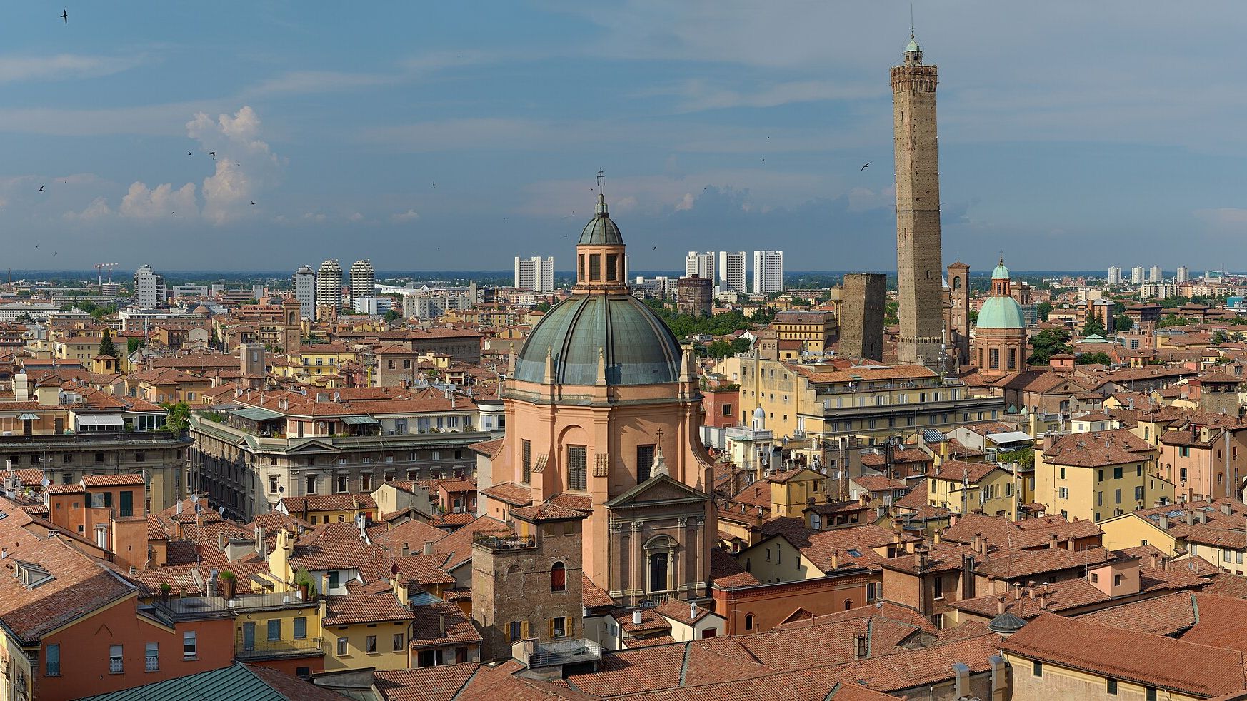 Terracotta rooftops of Bologna seen from the top of San Petronio, with the green dome of Santa Maria della Vita in the foreground and the tall Asinelli tower and leaning Garisenda behind