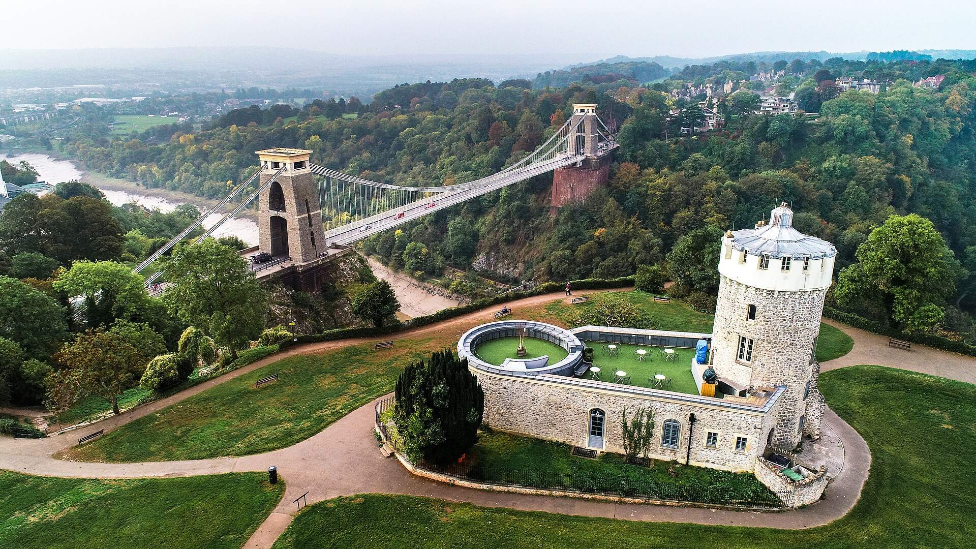 Aerial view of the Clifton Suspension Bridge spanning the wooded Avon Gorge in Bristol, with the stone tower of the Clifton Observatory in the foreground