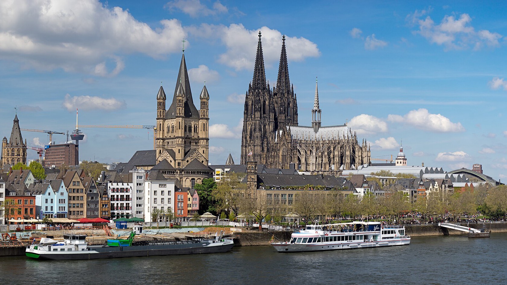 Cologne’s old town seen across the Rhine: gabled houses on the bank, the tower of Groß St. Martin and the twin spires of the cathedral under a blue sky with white clouds