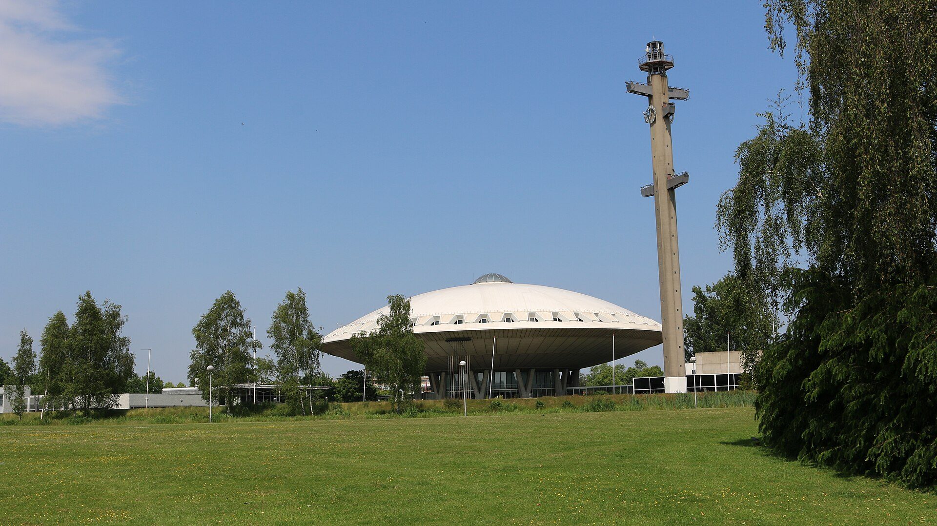 The Evoluon in Eindhoven, a white saucer-shaped building on V-shaped supports with its slender concrete tower beside it, seen across a meadow under a blue sky