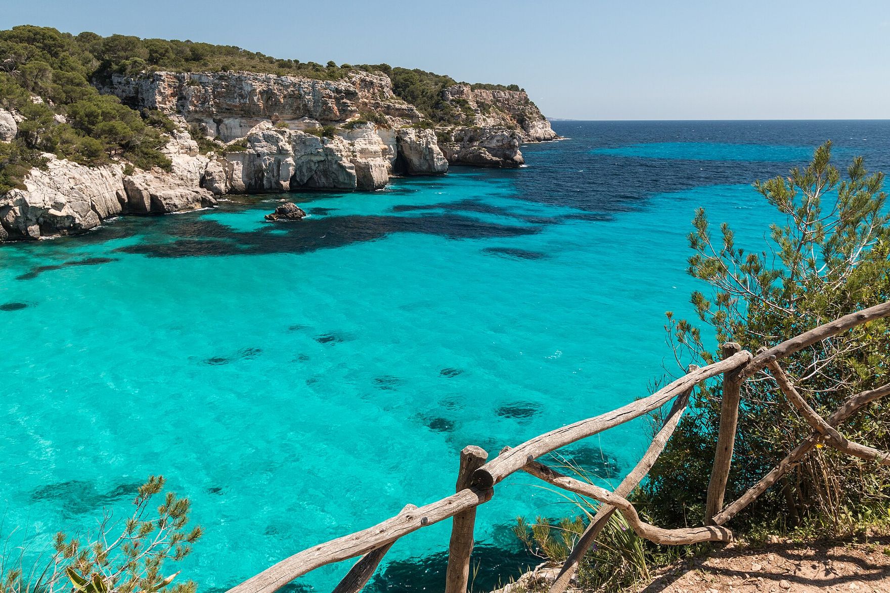 Pale limestone cliffs topped with pines above the clear turquoise water of Cala Macarella on Menorca, seen from a cliff path with a wooden fence