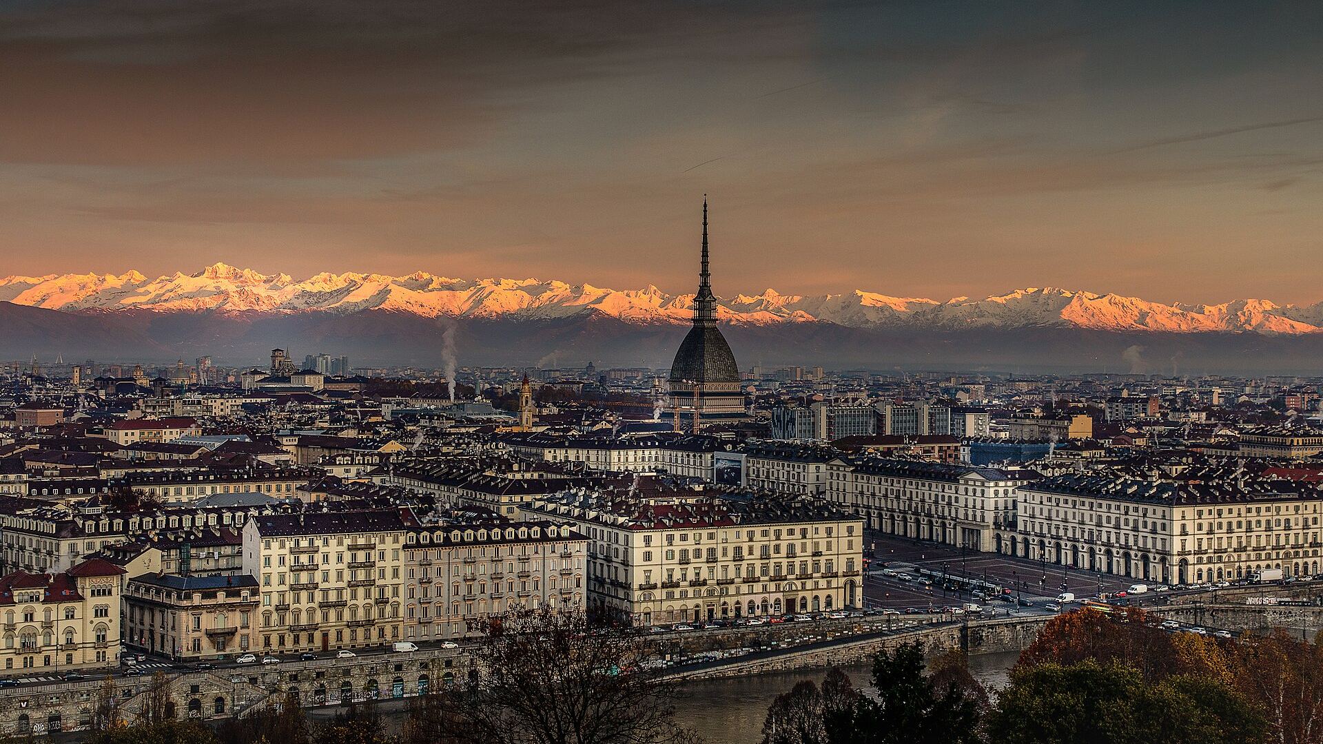 Turin at sunrise seen from Monte dei Cappuccini: the spire of the Mole Antonelliana above the rooftops, the arcades of Piazza Vittorio Veneto beside the Po, and the snow-covered Alps glowing pink behind